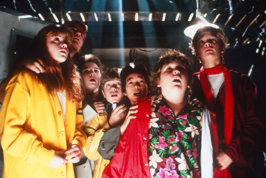 Photo of the Goonies cast, including Kerri Green, Josh Brolin, Corey Feldman, Sean Astin, Ke Huy Quan, Jeff Cohen, and Martha Plimpton, looking up in wonder at a bright light from above.