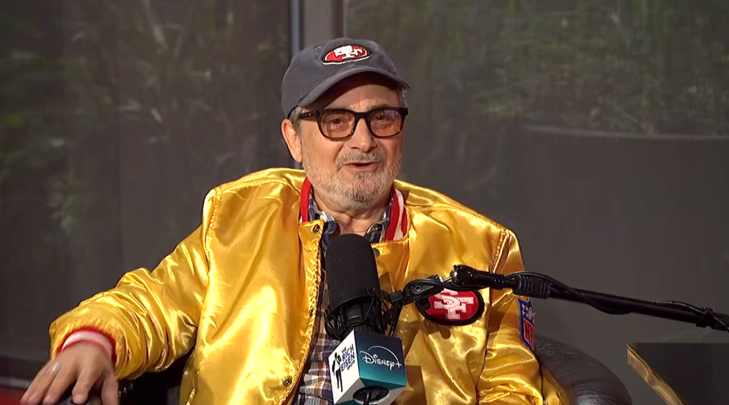 Actor Kevin Pollak wearing a San Francisco 49ers hat and jacket, speaking into a microphone.