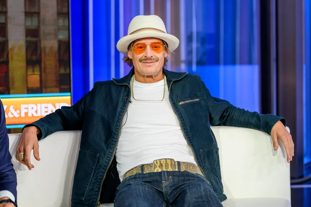 Kid Rock sitting on a white sofa during a television appearance, wearing a white hat and orange-tinted sunglasses.