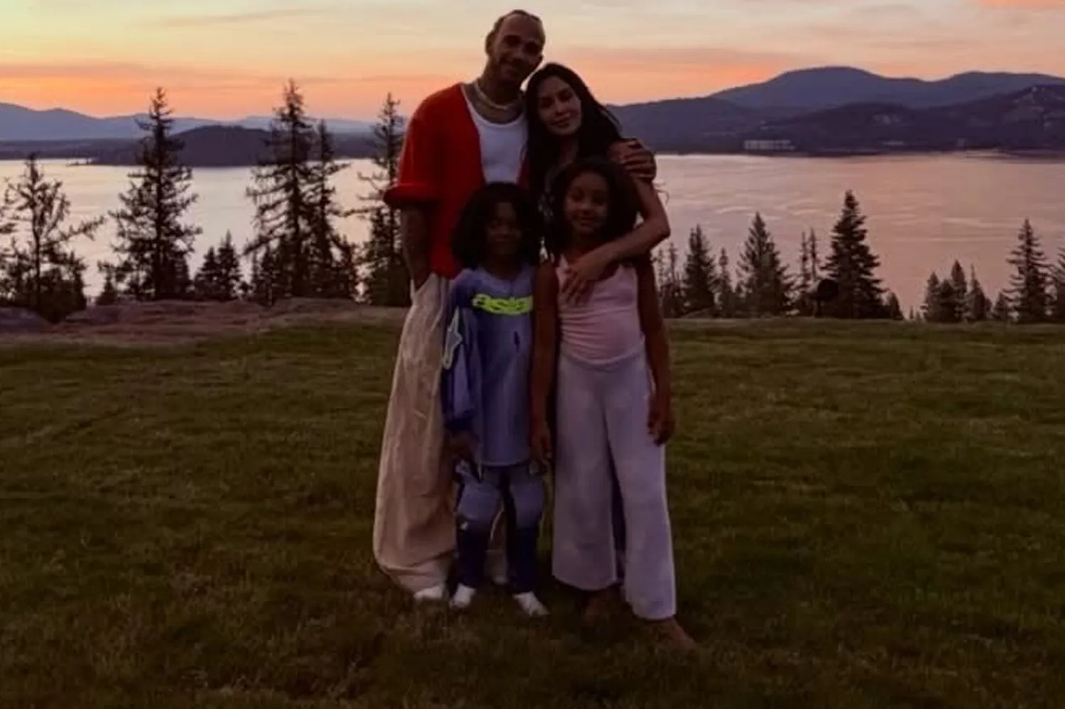 Kim Kardashian and Lewis Hamilton pose together with her kids outdoors at sunset near a lake.
