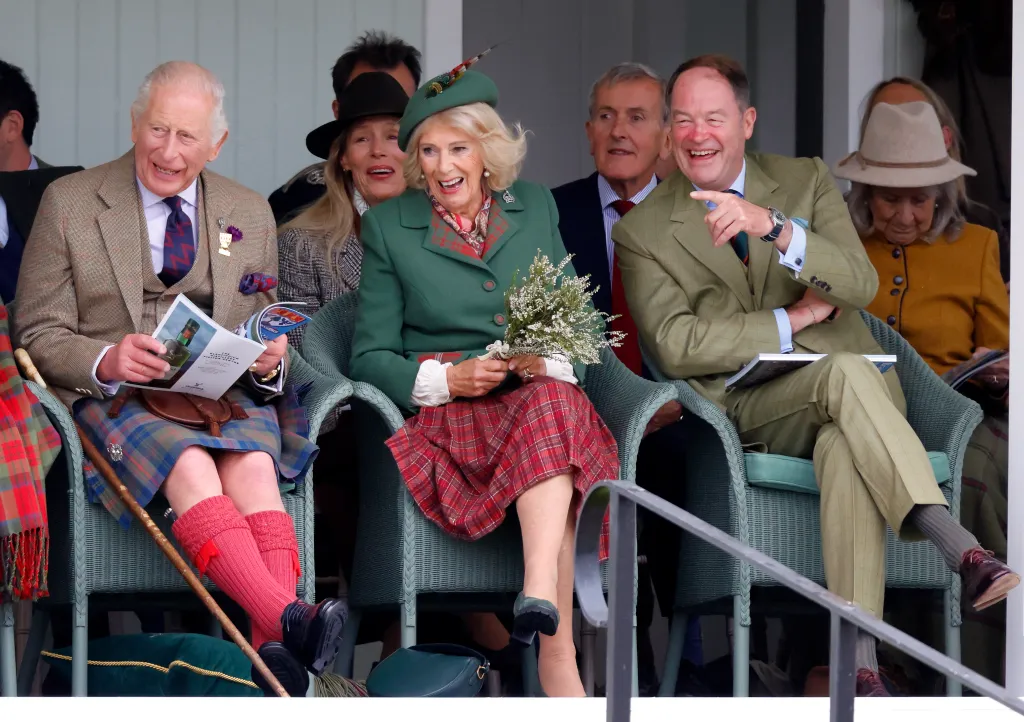 King Charles III, ub a brown blazer and brown and blue checked kilt, with red socks, and blue shirt, Queen Camilla, in a green jacket and red kilt and green hat, and General Sir Patrick Sanders, in a green suit, laughing while seated outdoors.