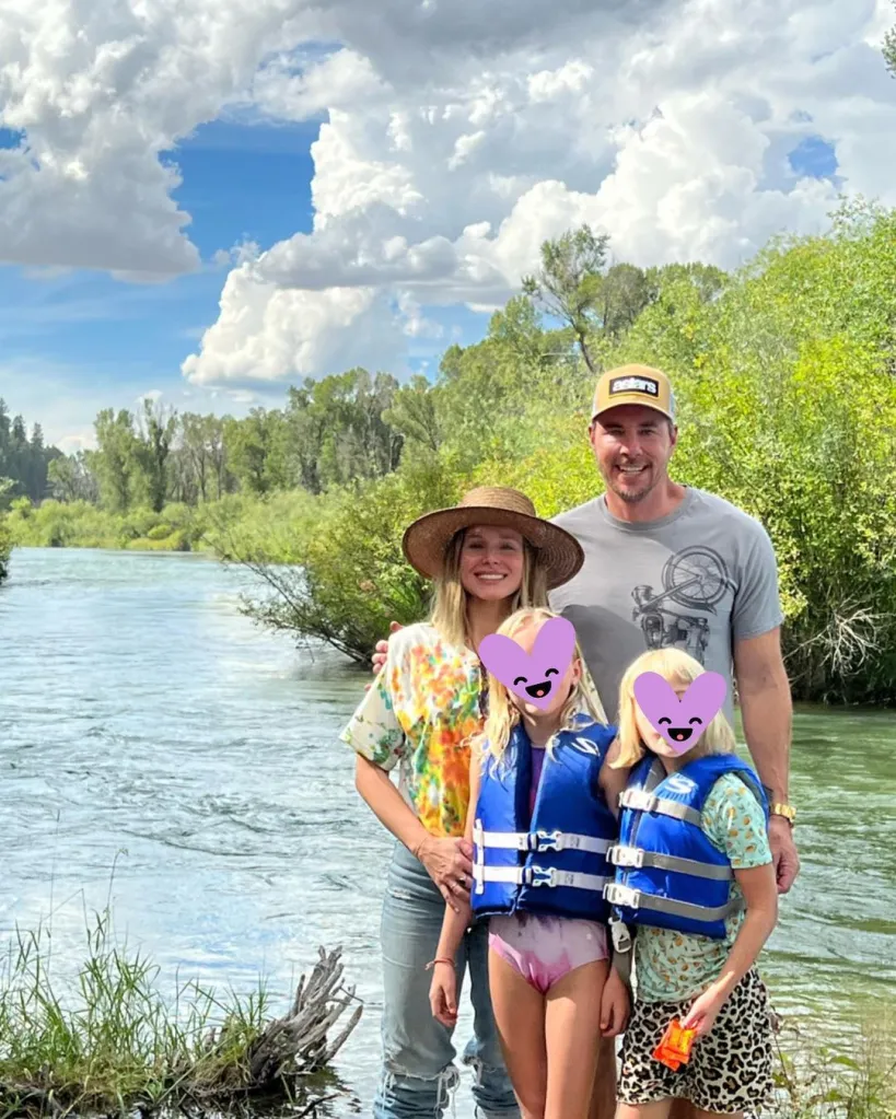 Kristin Bell and Dax Shepard pose outdoors with their two daughters by a river on a sunny day, with the daughters' faces covered by purple heart emojis.