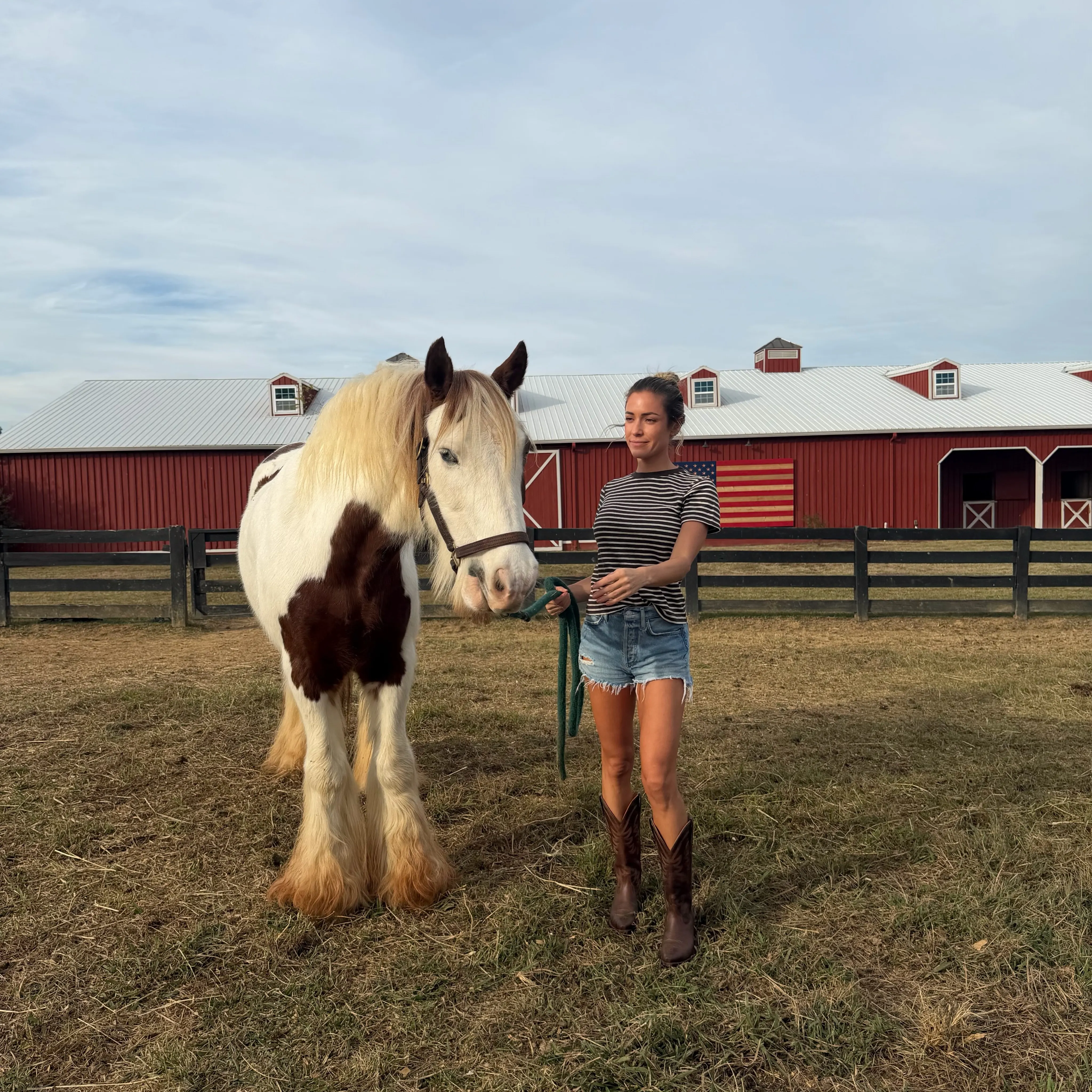 Kristin Cavallari wearing shorts and boots, stands in a pasture holding the reins of a piebald horse in front of a red barn.