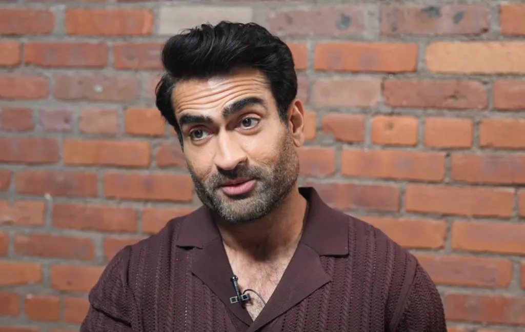 Kumail Nanjiani in a brown shirt standing against a brick wall.