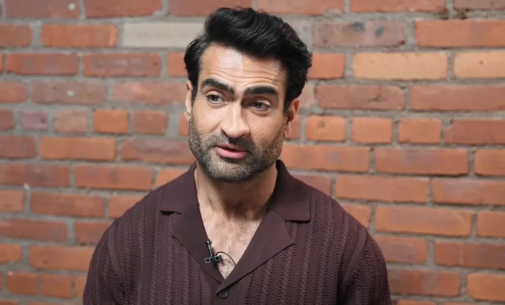 Kumail Nanjiani in front of a brick wall.