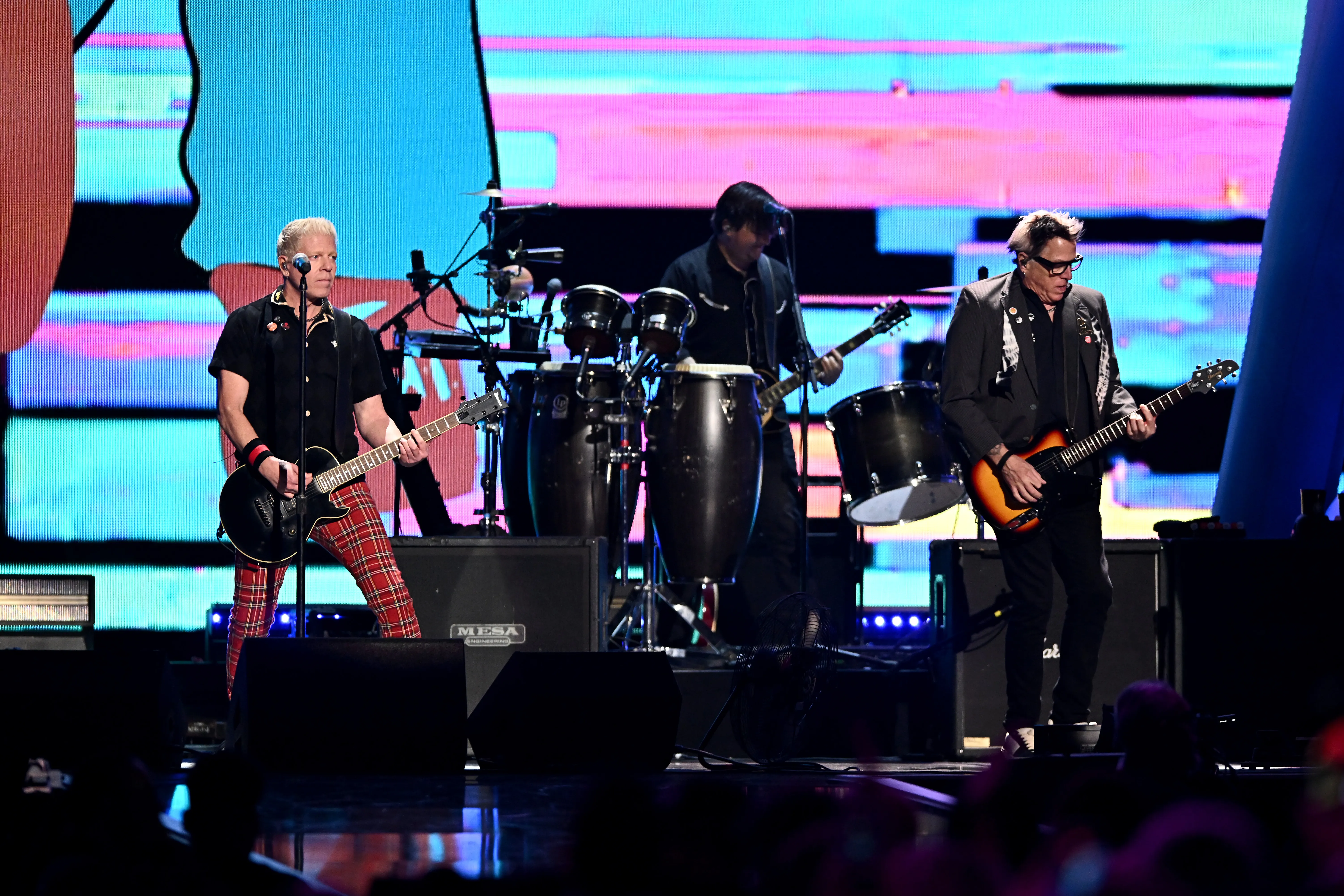Dexter Holland and Noodles of The Offspring perform onstage, with Holland on the left playing a black guitar and wearing red plaid pants, and Noodles on the right playing a sunburst guitar and wearing a dark suit jacket and glasses.