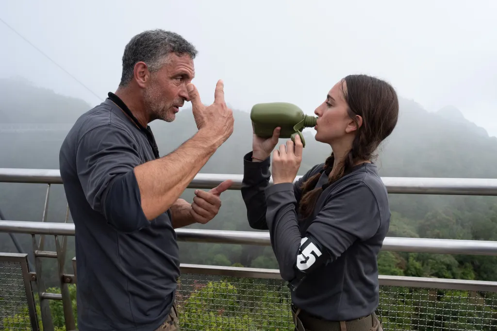 Mauricio Umansky and Alexia Umansky stand on a foggy outdoor platform, with Mauricio pointing at his face as Alexia drinks from a green canteen.