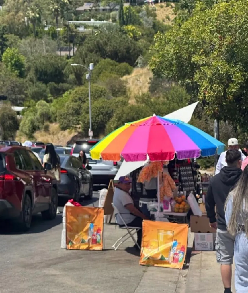 A street vendor with a rainbow umbrella surrounded by heavy traffic near the Hollywood sign.