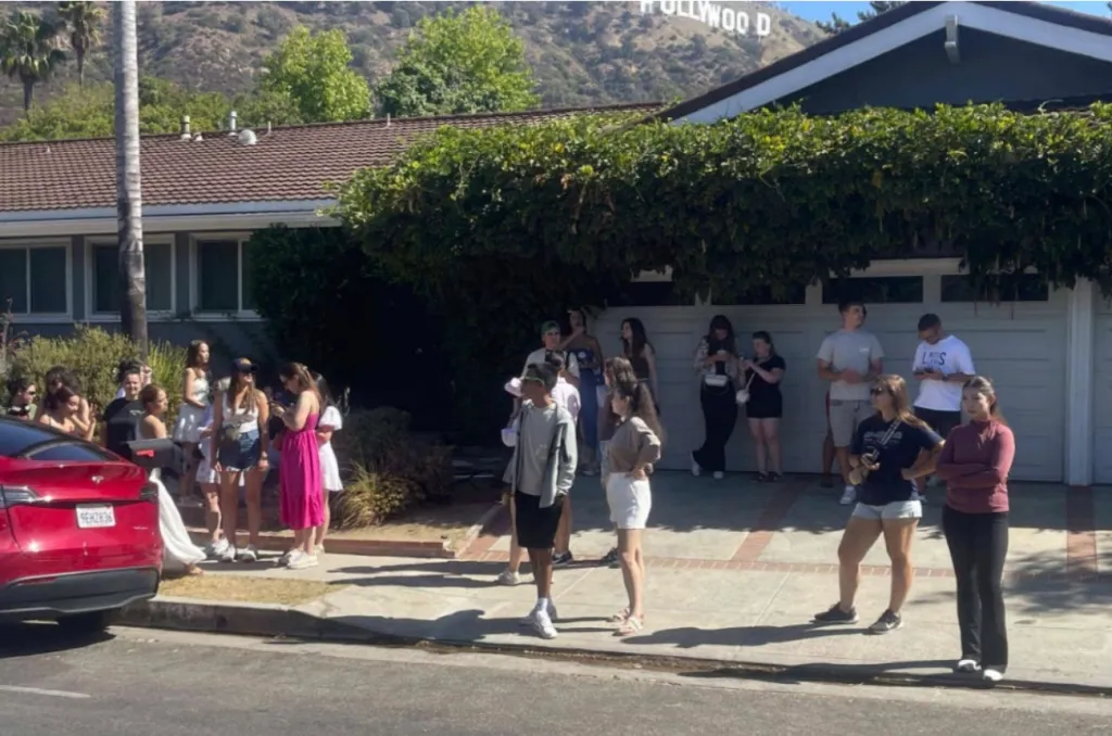 Visitors and onlookers gather on the street and driveway of a residential home near the Hollywood sign.
