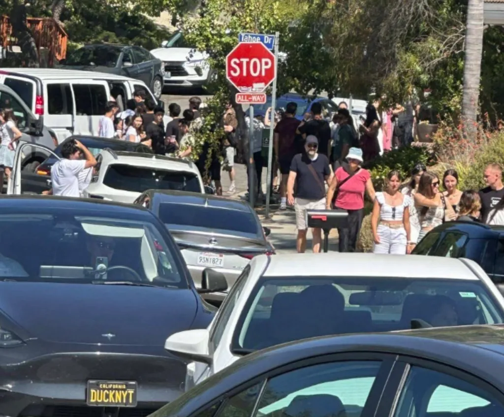 Crowded residential street near the Hollywood sign, congested with parked and stopped cars and pedestrians walking on the sidewalk.