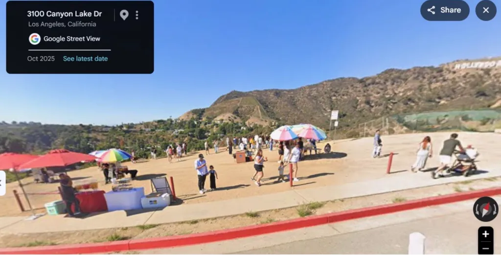 Google Street View of a crowded viewing area near the Hollywood sign in Los Angeles, showing vendors and visitors gathered on a dirt lot.