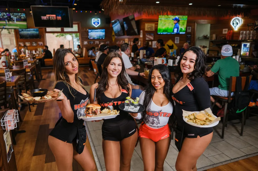 Four Hooters servers pose in a restaurant, holding plates of food and drinks.