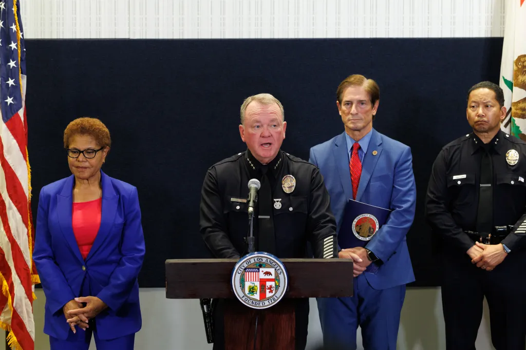 LAPD Police Chief Jim McDonnell speaks at a press conference, flanked by officials and American and California flags.