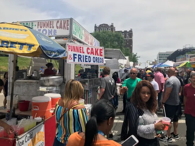 A crowded street fair featuring food booths selling funnel cakes, fried Oreos, and lemonade.