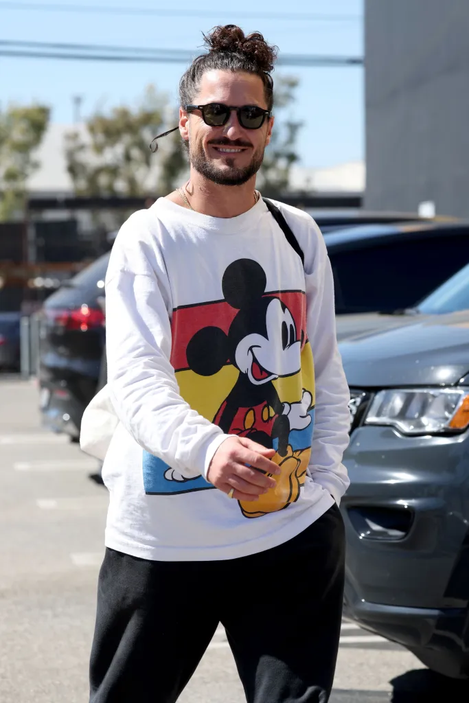 Val Chmerkovskiy smiling while wearing a Mickey Mouse sweatshirt and sunglasses, walking in a parking lot.