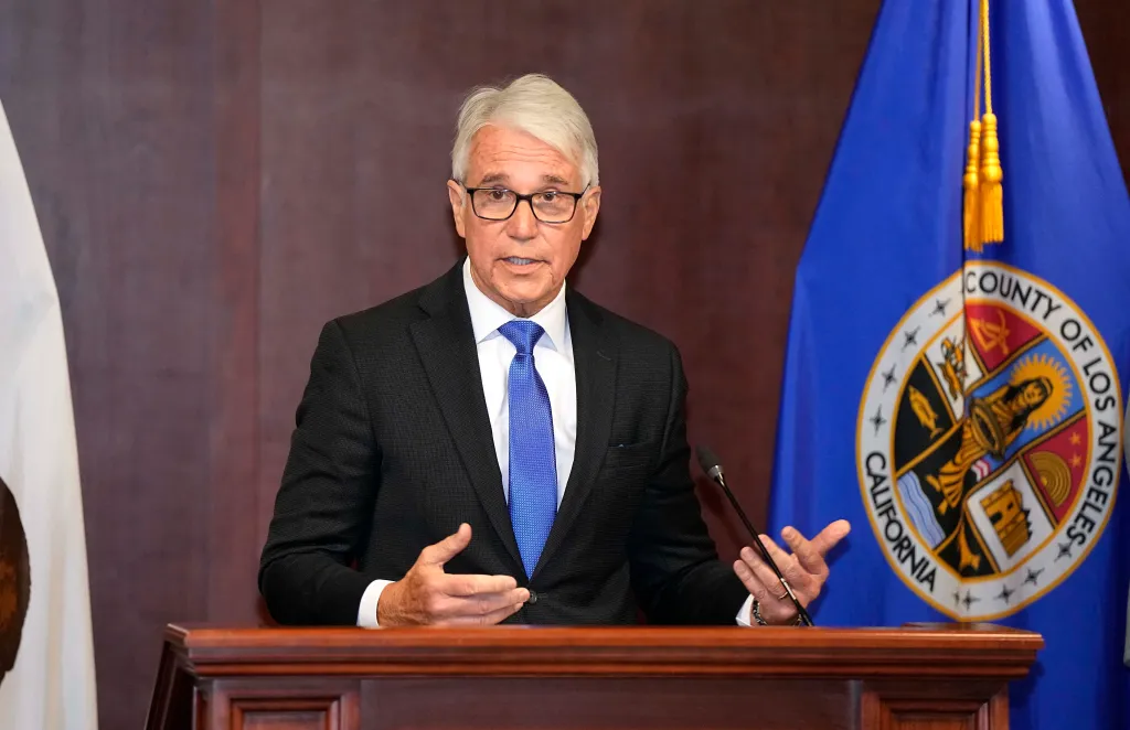 Los Angeles County District Attorney George Gascón speaks at a podium during a news conference, with the Los Angeles County seal visible on a flag in the background.