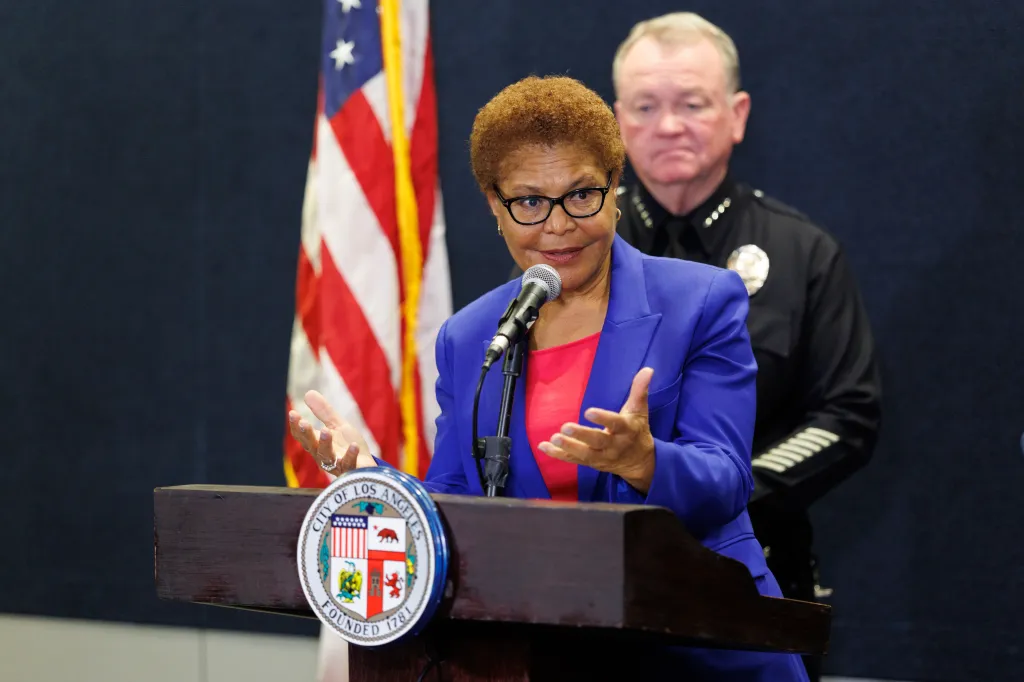 Los Angeles Mayor Karen Bass speaks at a press conference at LAPD Headquarters, with an officer standing behind her.