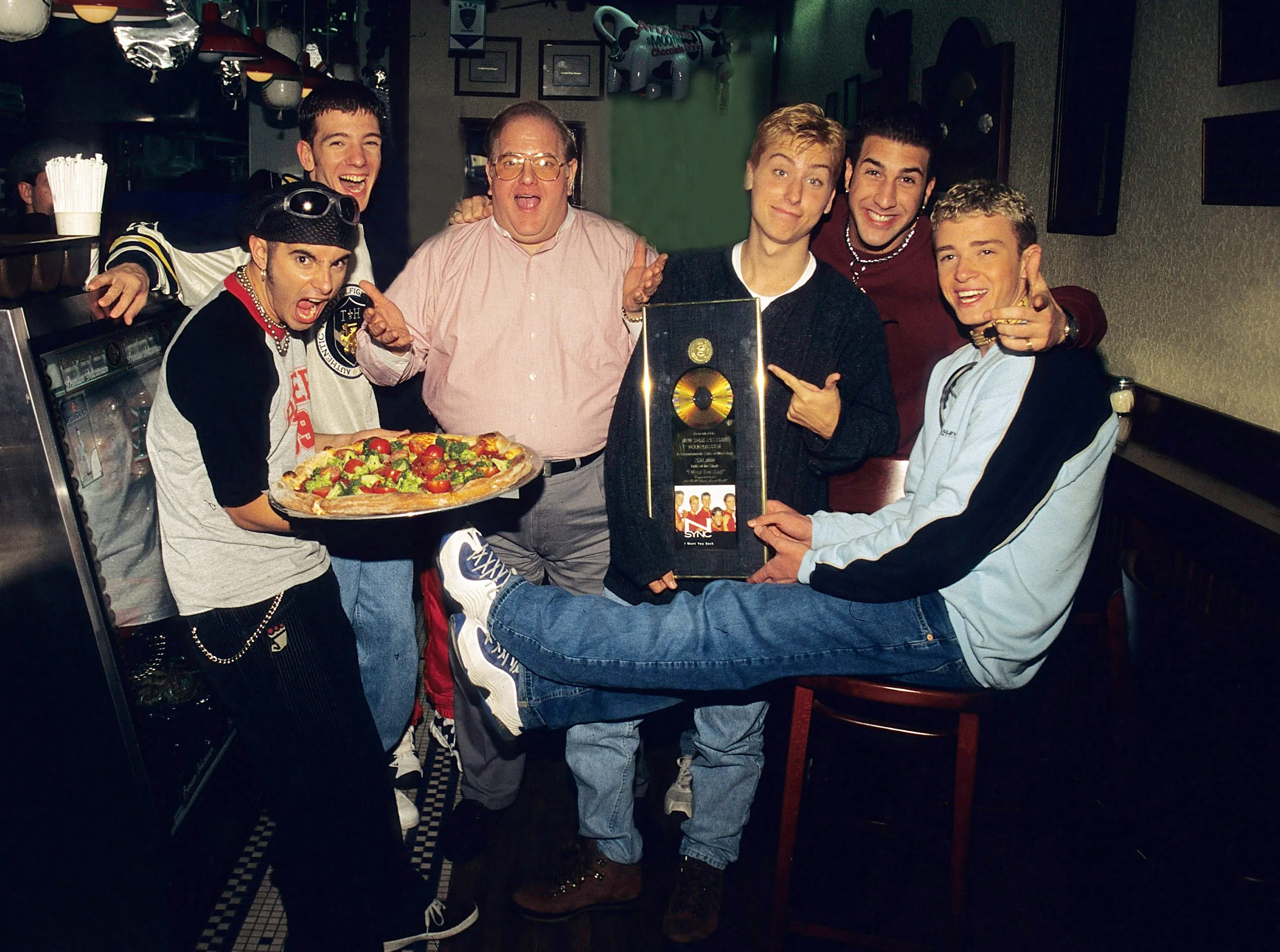 Lou Pearlman poses with the members of N'Sync—Chris Kirkpatrick, JC Chasez, Lance Bass, Joey Fatone, and Justin Timberlake—while holding a pizza and a gold record award in a pizza restaurant.