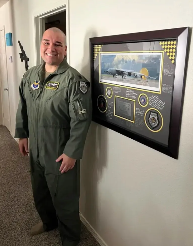 Lt. Col. Gabriel 'Journee' Estrella standing smiling in his flight suit next to a framed memorial display.