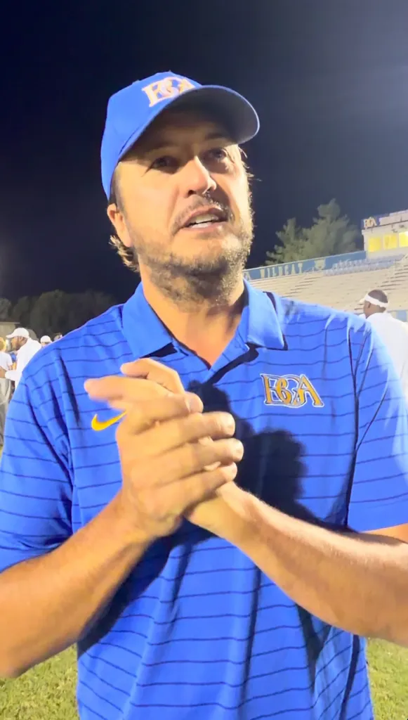 Luke Bryan wearing a blue polo shirt and matching baseball cap while clasping his hands together at a football game.