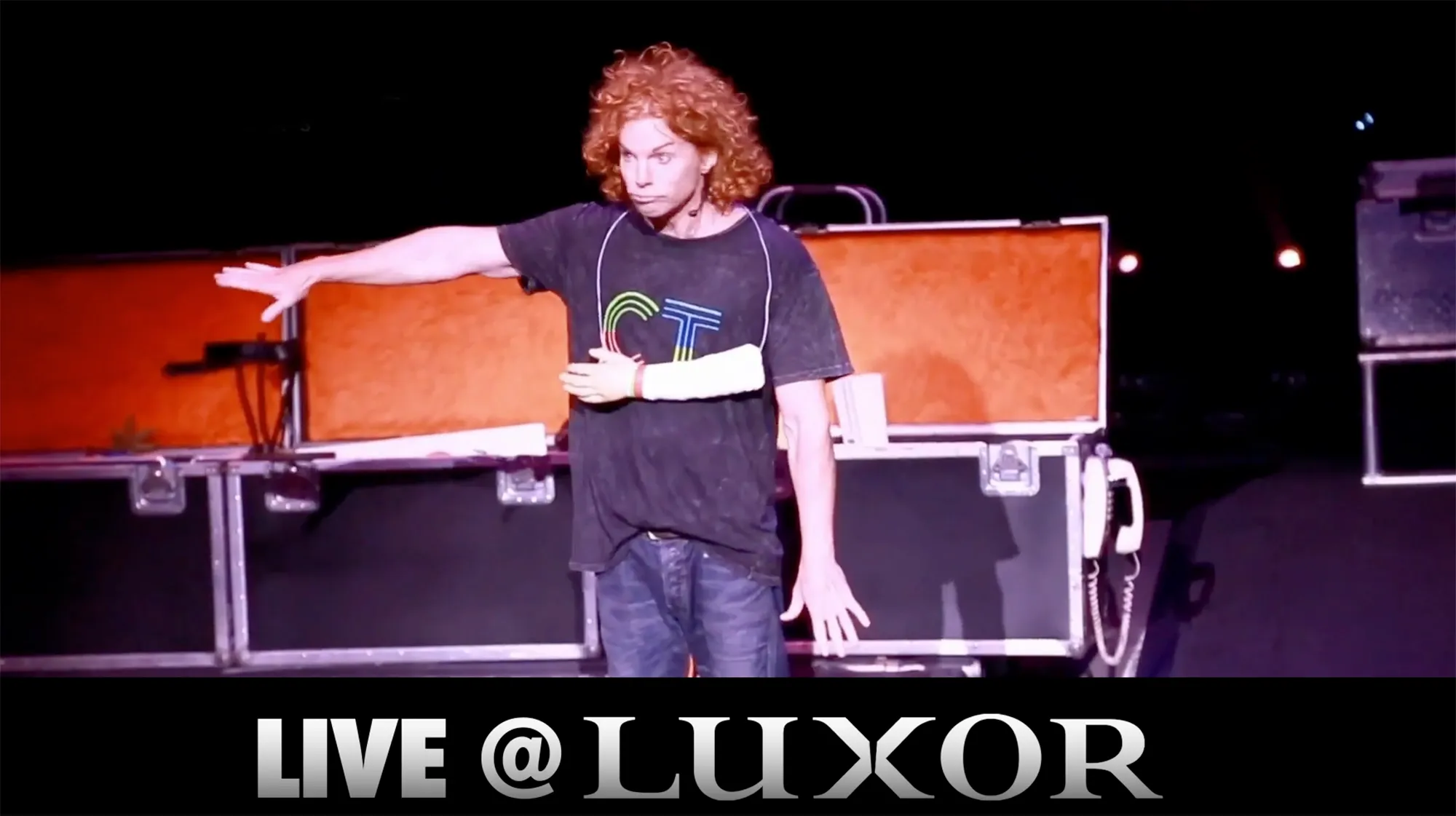 Comedian Carrot Top performs on stage with an open prop case behind him and the text
