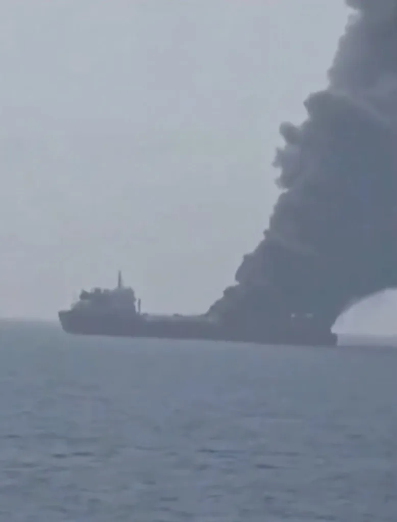 A large ship on fire with thick black smoke rising into the sky in the Strait of Hormuz.