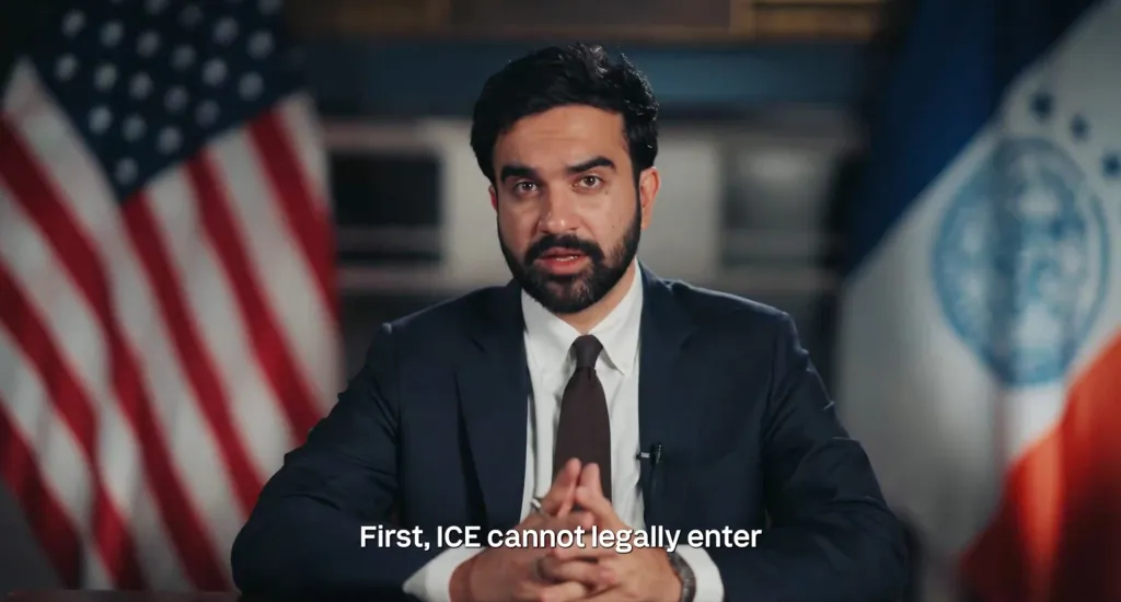 Zohran Mamdani speaking in a video with American and New York City flags in the background.