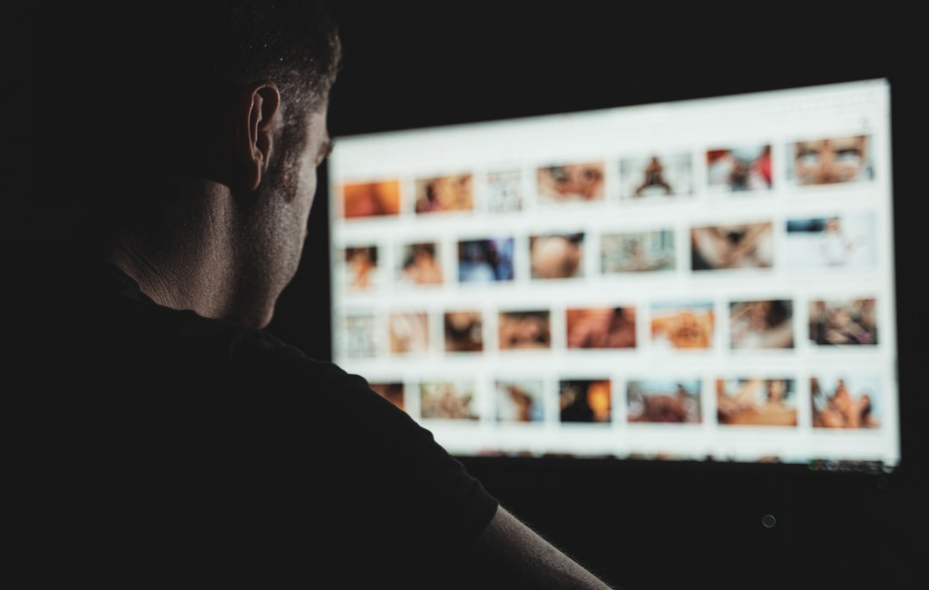A man browses an adult website on a computer screen in a dark room.