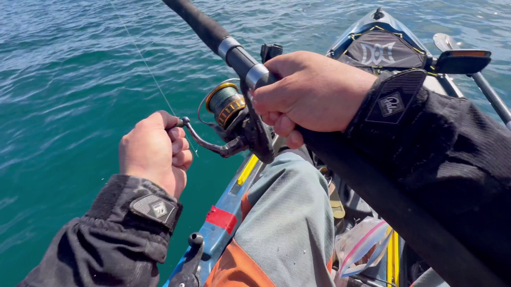 First-person view of a man holding a fishing rod and reel while sitting in a kayak on the ocean.