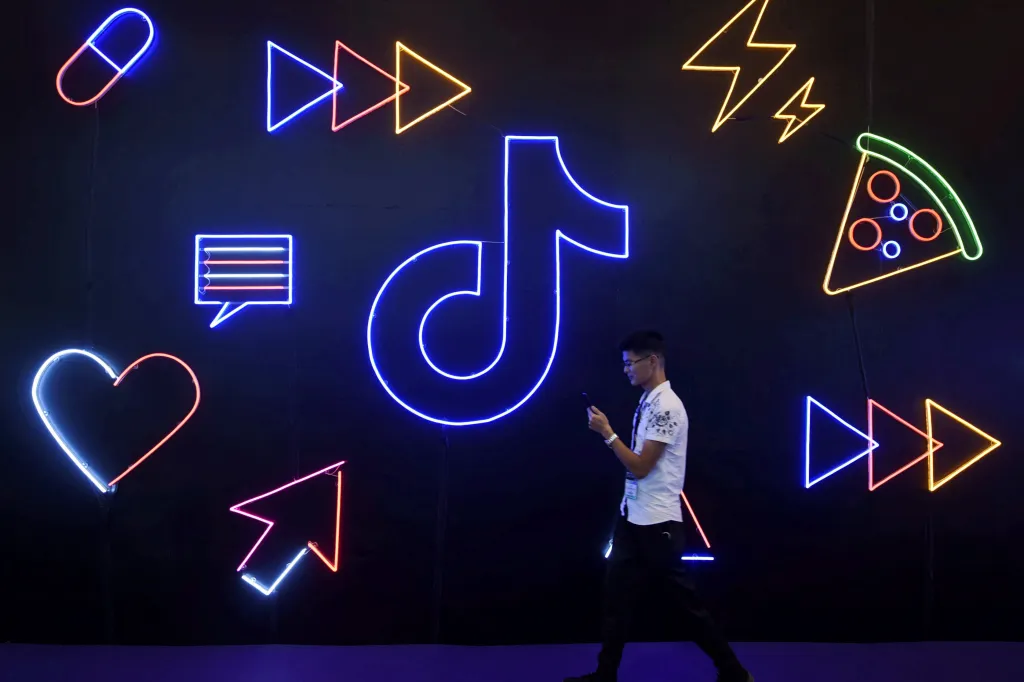 A man holding a phone walks past a large neon sign of ByteDance's app TikTok, surrounded by other neon signs.
