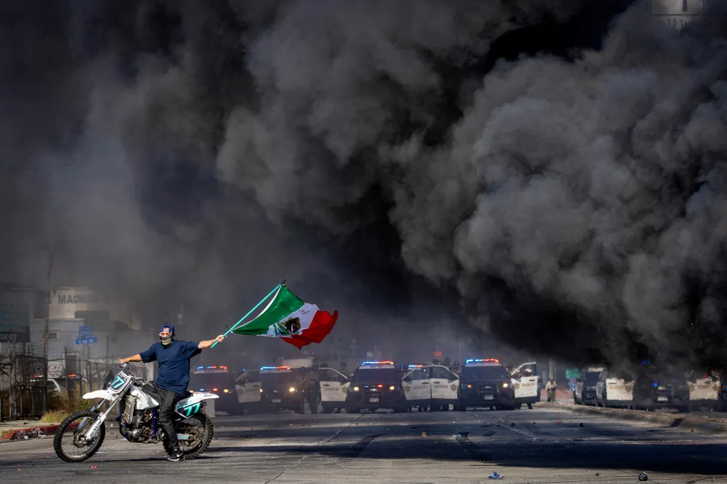 A man on a motorcycle waves a Mexican flag during a standoff between protesters and law enforcement as thick black smoke billows into the air in front of a line of police cruisers.