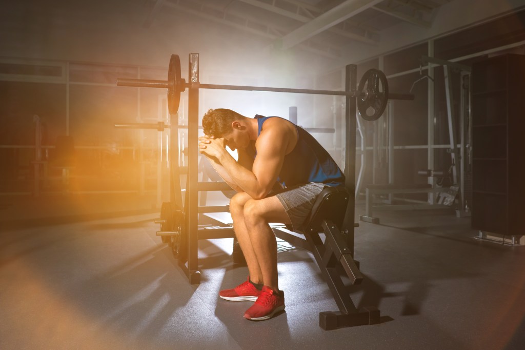 A tired athlete sits hunched over on a gym bench, holding his head in his hands.