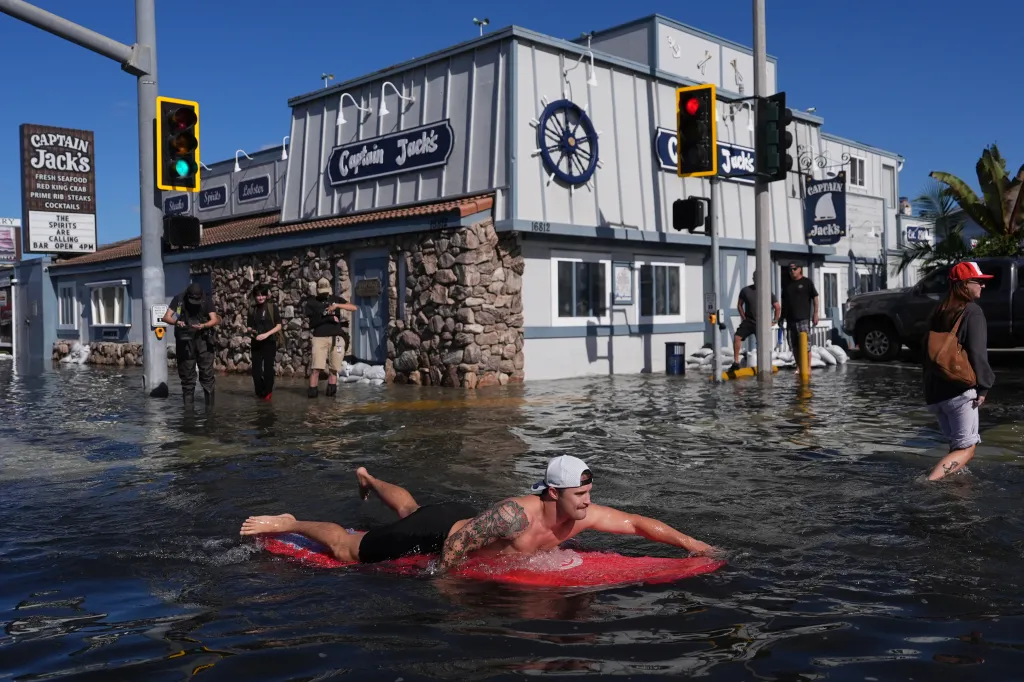 A man paddles a red surfboard through floodwaters in front of a restaurant in Huntington Beach, California.
