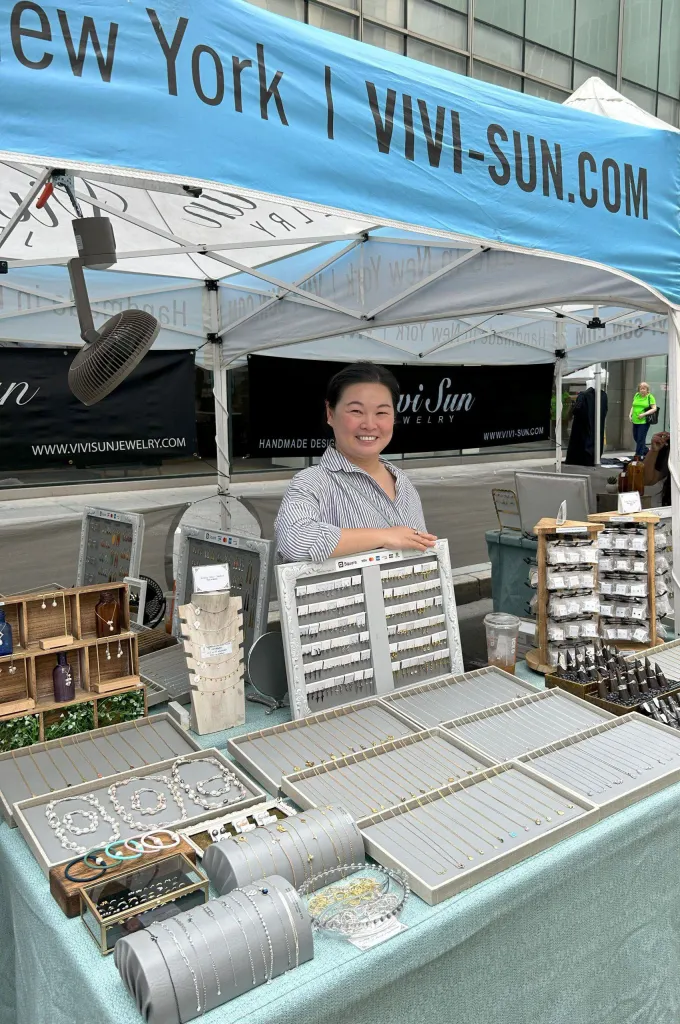 A smiling vendor stands behind a table displaying handmade jewelry, including necklaces, earrings, and bracelets, at a street fair booth for Vivi-Sun.