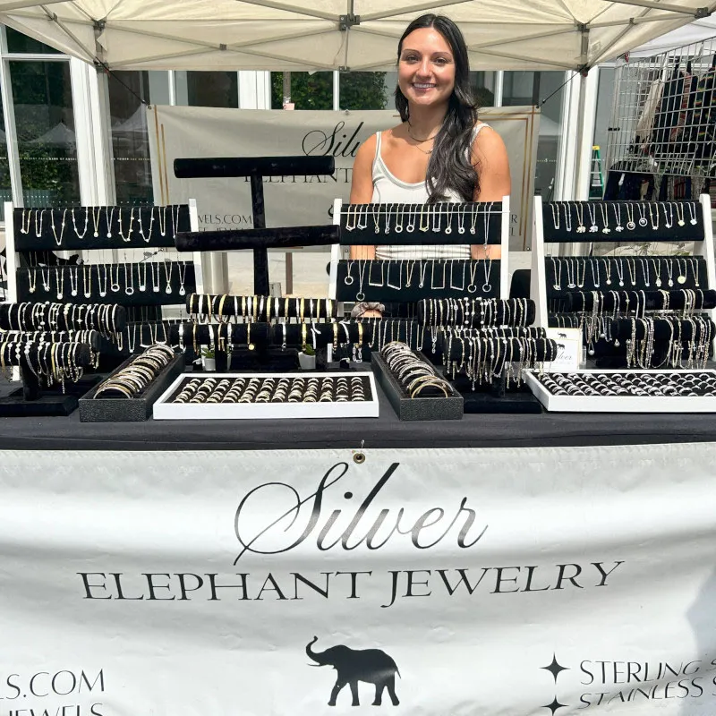 A smiling vendor stands behind a table displaying necklaces, bracelets, and rings at an outdoor street fair booth for Silver Elephant Jewelry.