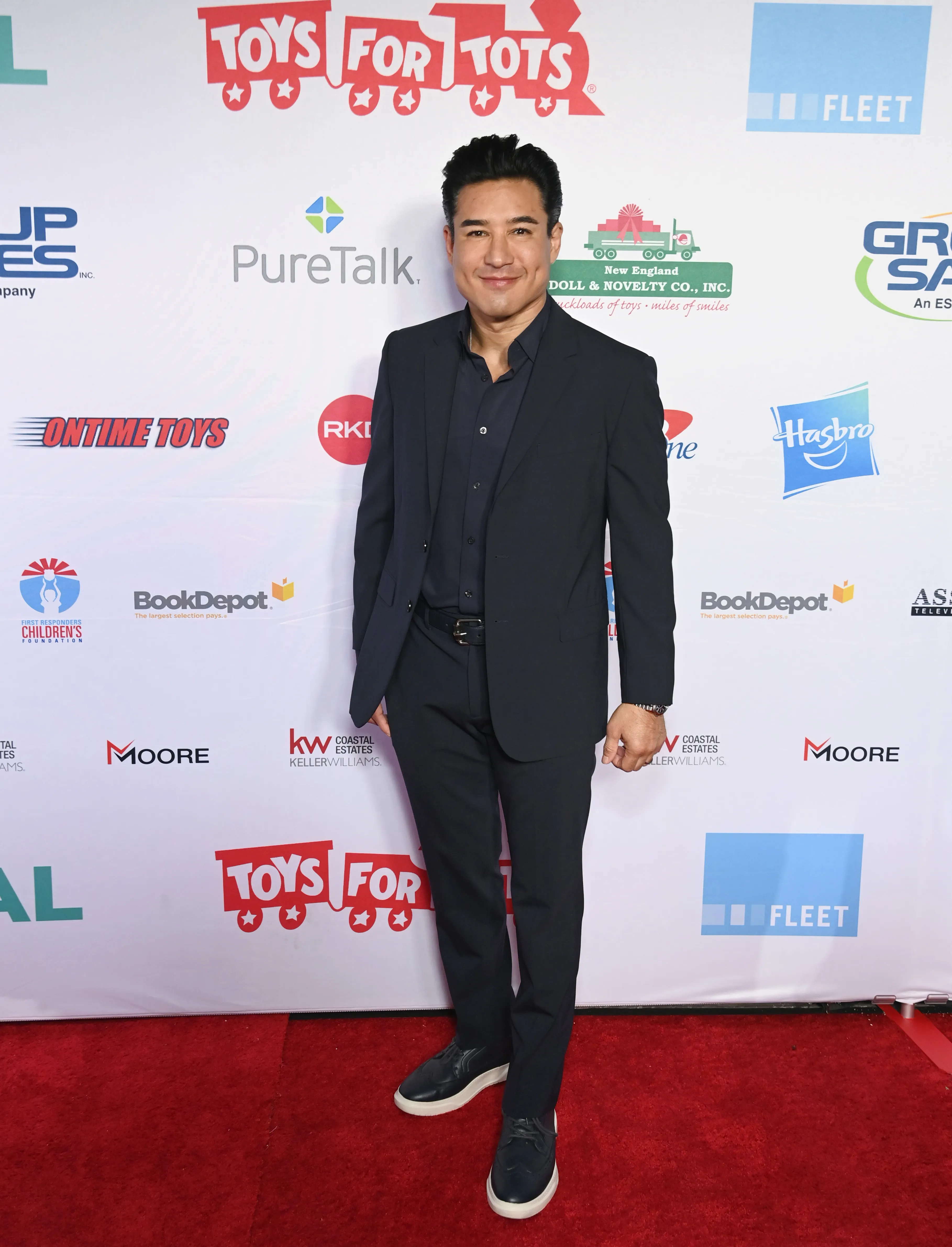 Mario Lopez poses in a dark suit on a red carpet in front of a sponsor backdrop.