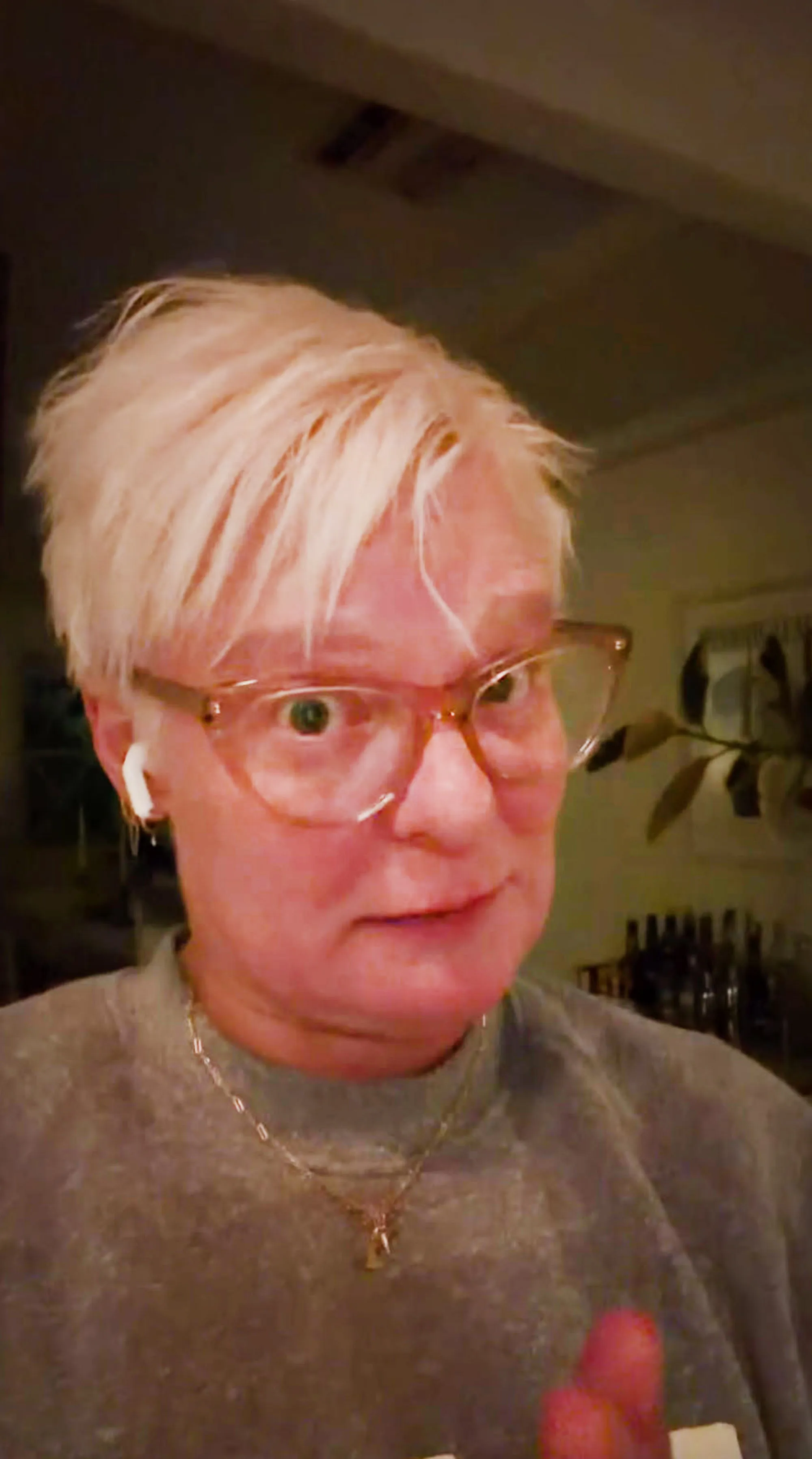 Martha Plimpton wearing glasses and a grey sweatshirt, with short blonde hair and an earbud.