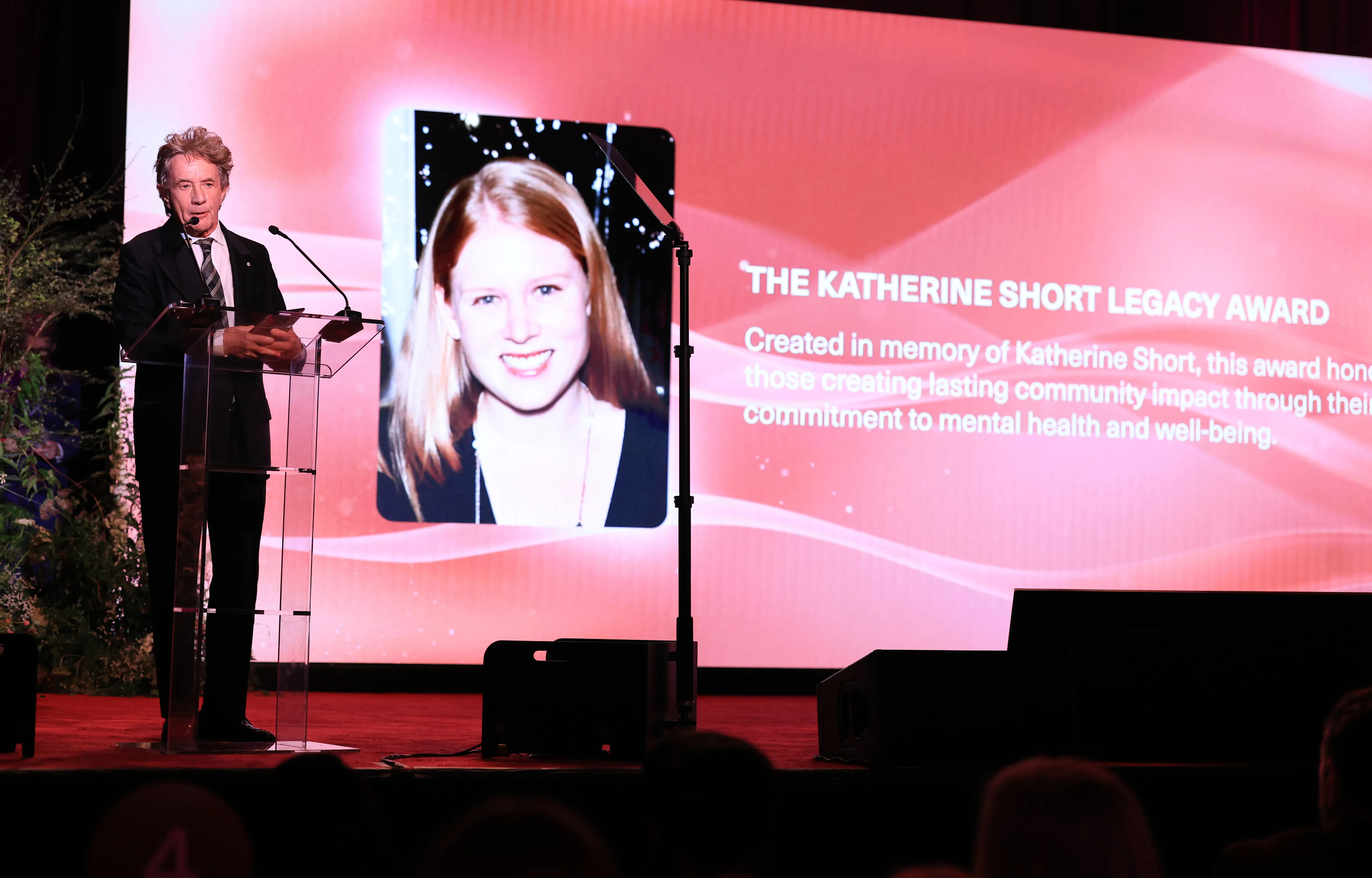 Martin Short speaks at a podium in front of a screen displaying a photo of Katherine Short and text about The Katherine Short Legacy Award.