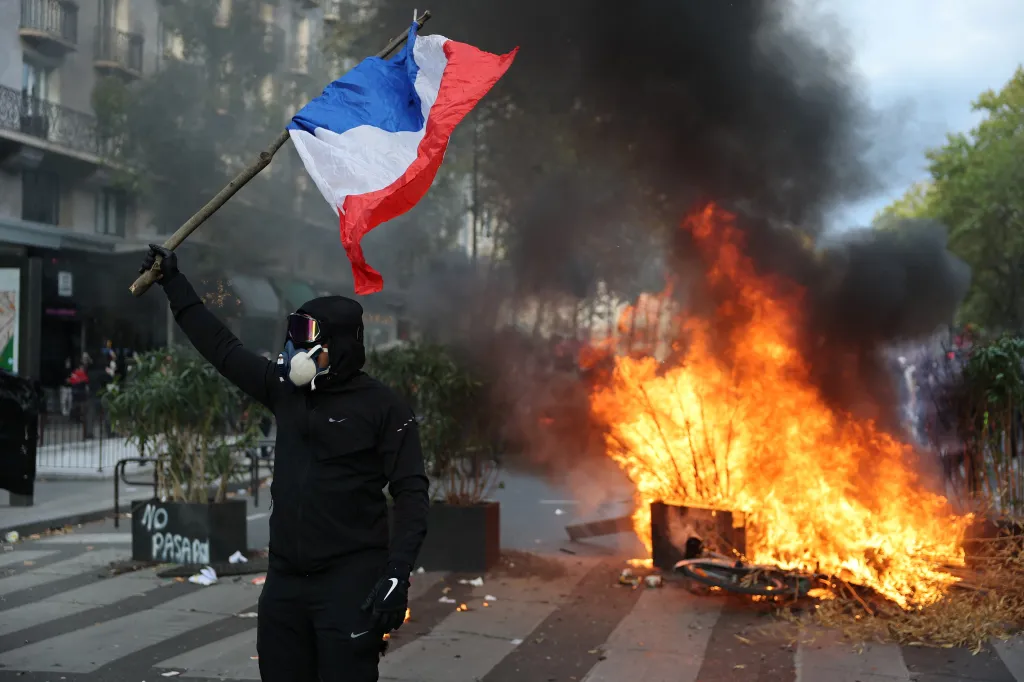 A masked protester holding a French flag in front of a burning barricade during a demonstration in Paris.