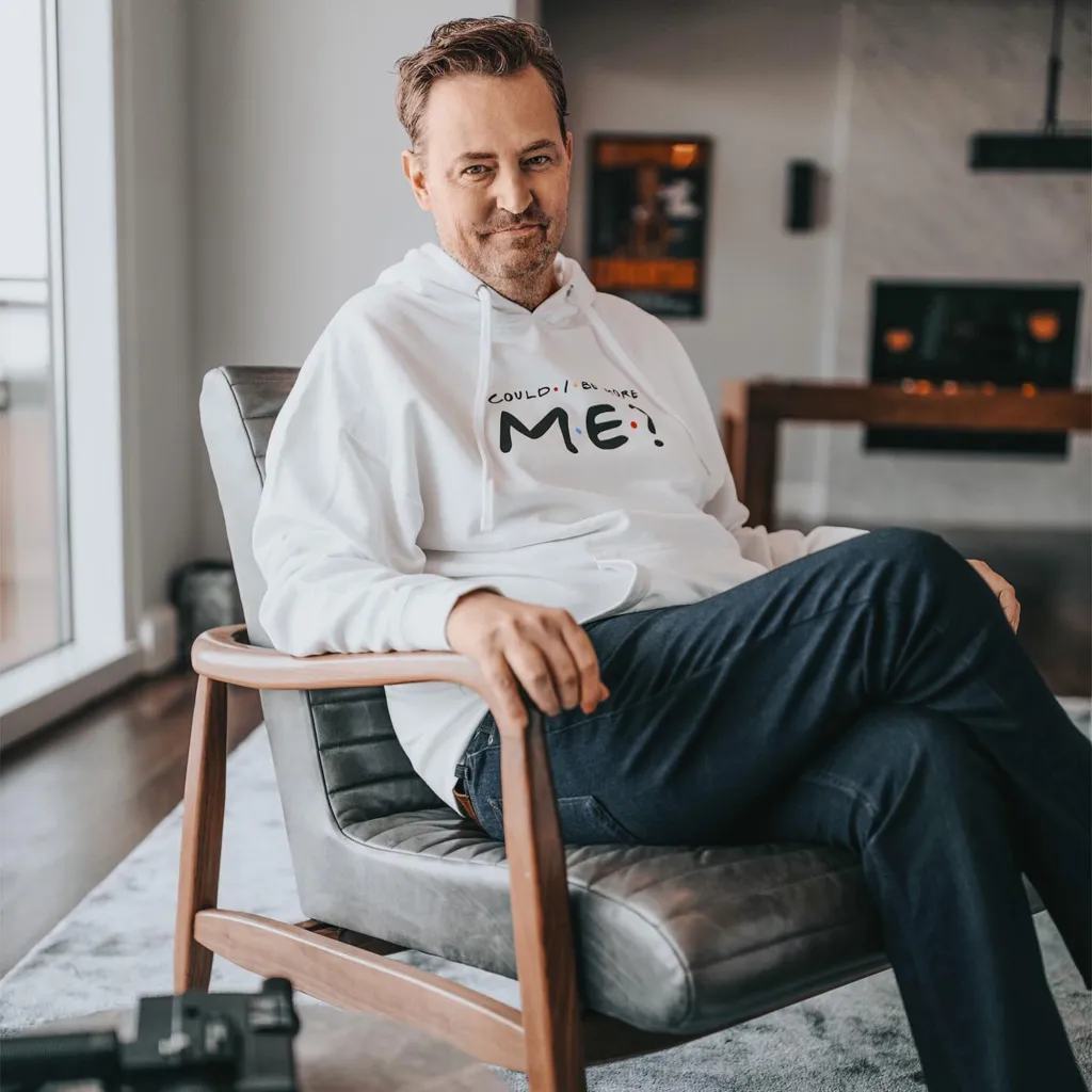 Matthew Perry sitting in an armchair while wearing a white hoodie that reads 