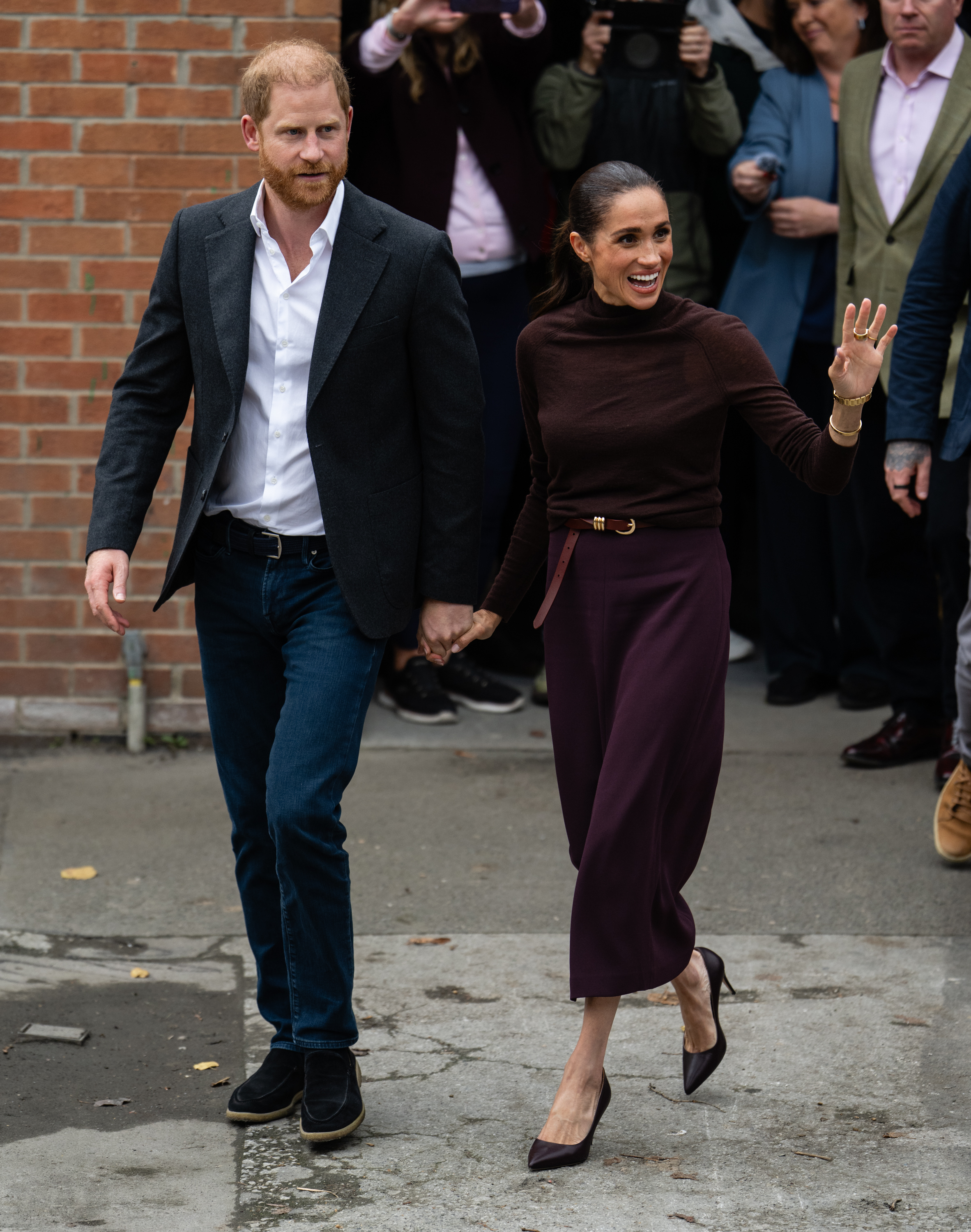 Prince Harry and Meghan, Duchess of Sussex, holding hands and walking past a brick wall.