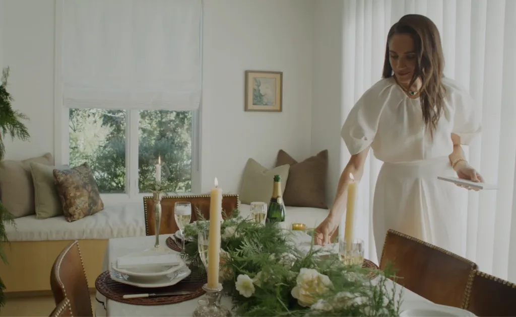 Meghan Markle wearing a white dress sets a dining table decorated with candles, champagne, and floral arrangements.