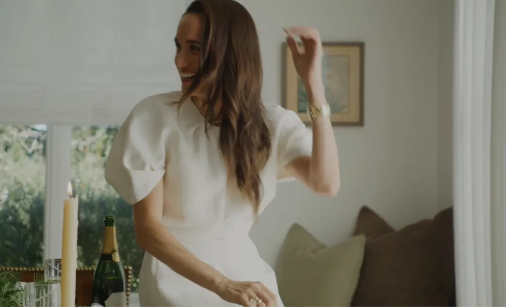 Meghan Markle smiling while wearing a white dress in an indoor setting.
