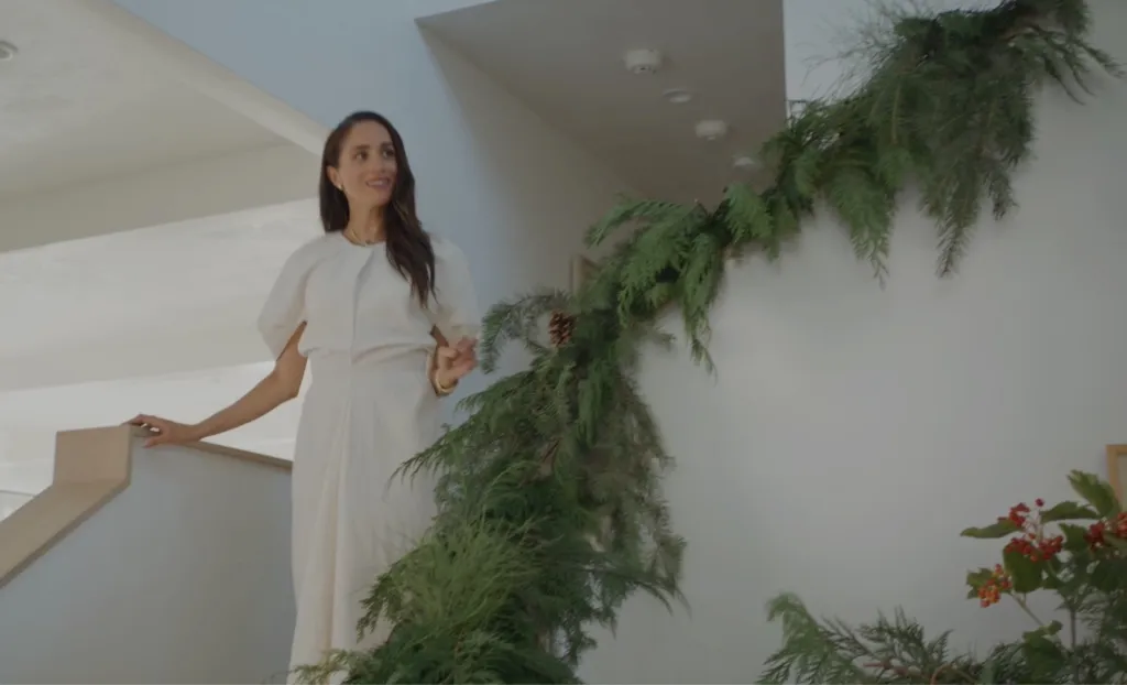 Meghan Markle wearing a white dress standing on a staircase adorned with pine greenery.