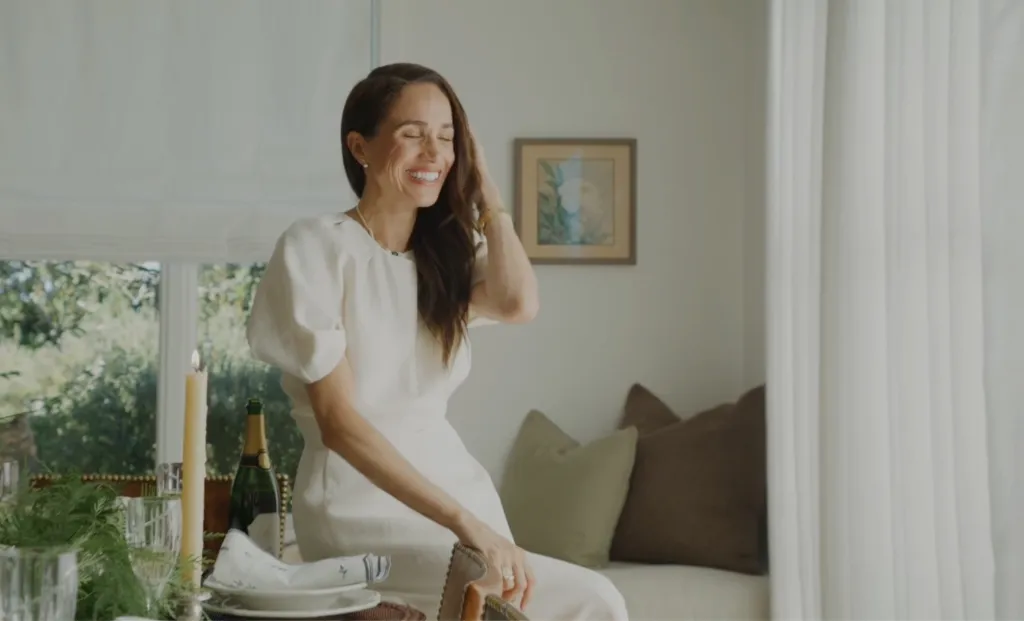 Meghan Markle smiling while wearing a white dress in a brightly lit room.