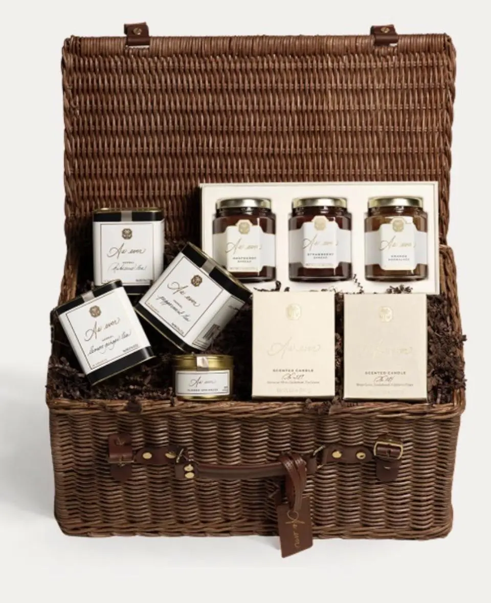 An open wicker hamper filled with Meghan Markle's As Ever holiday gift items, including jars of spread, tea tins, and candles.