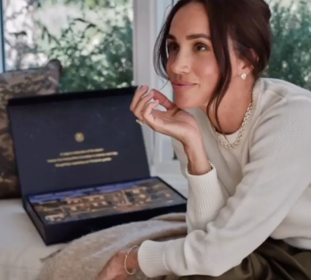 Meghan Markle, in a cream top, smiling with her hand near her chin next to an open black box.