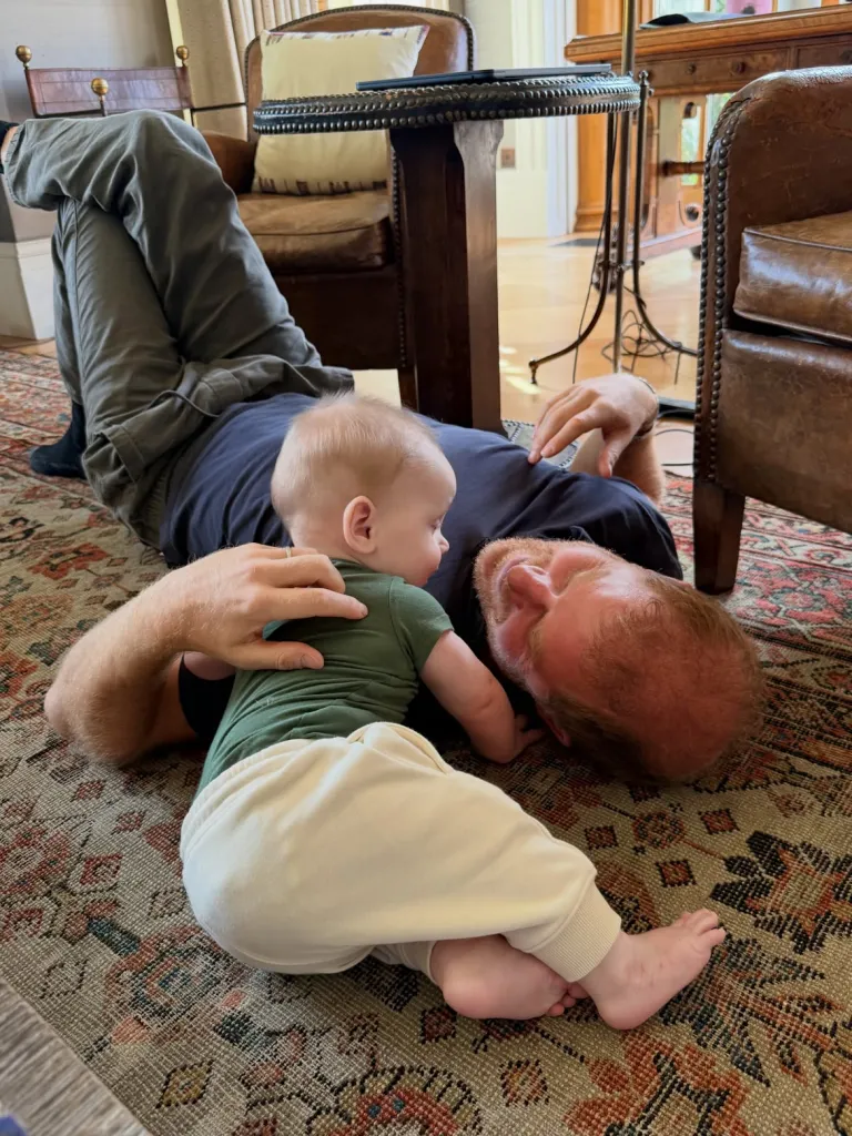 Prince Harry in a blue T-shrit and black pants, lying on the floor playing with baby Jack.
