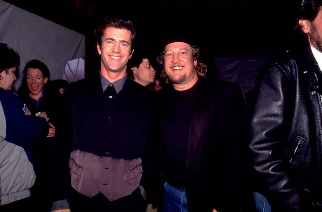 Mel Gibson and John Anderson smiling together in a dimly lit room.