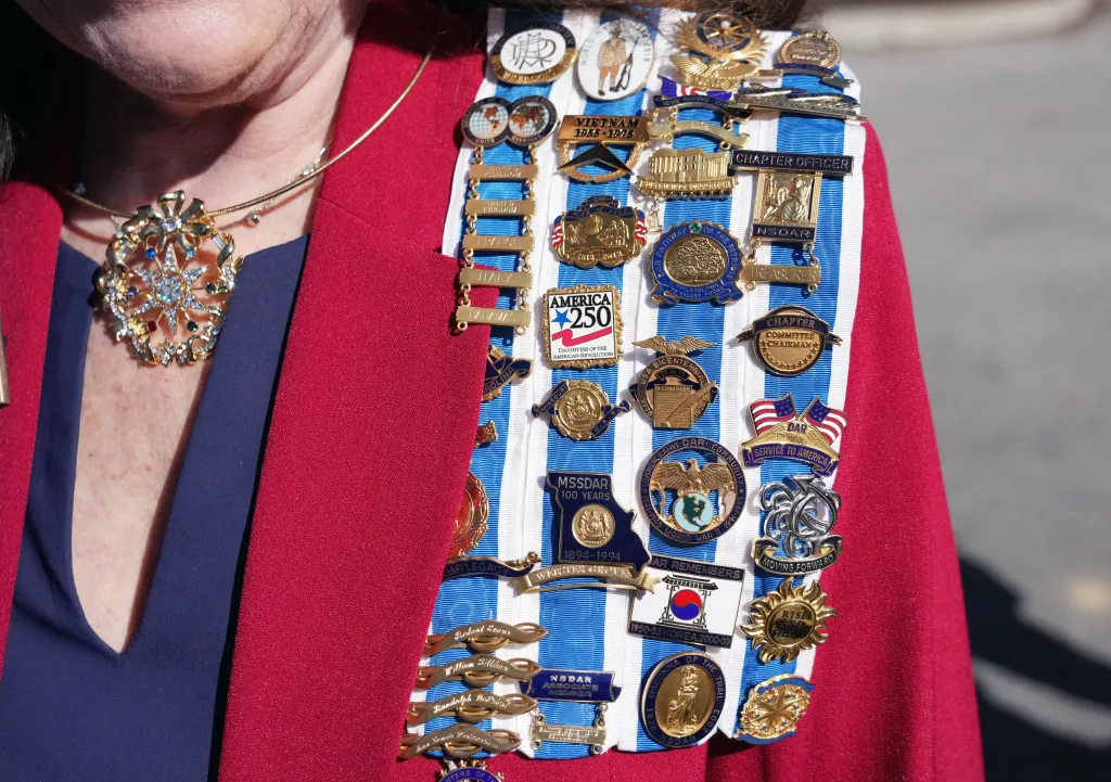 A member of the Daughters of the American Revolution wears her medals on a blue and white ribbon attached to her red jacket.